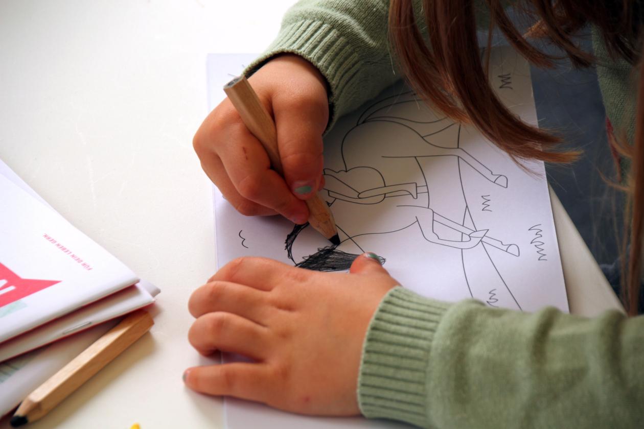 Kind malt mit Buntstift eine Figur auf einem Blatt Papier aus.